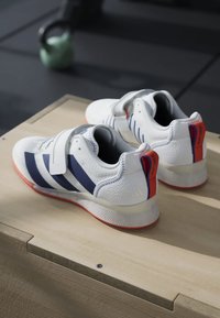Hvide atletiske sko med navy striber, designet med Velcro-lukninger, tekstureret overdel og røde detaljer på hælen og sålen.