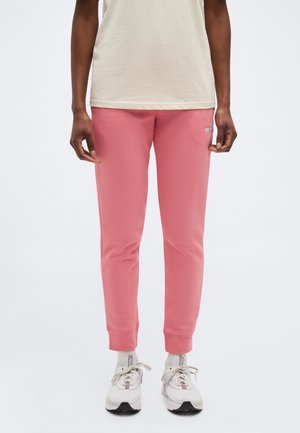 Ember világos rózsaszín melegítőnadrágot, fehér zoknit, fehér tornacipőt és bézs rövid ujjú felsőt visel, egyszínű háttér előtt áll.