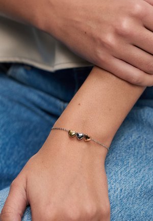 Zarter Silberkettenarmband mit drei Herzanhängern in Gold, Silber und Roségold, getragen am Handgelenk über blauer Jeans.