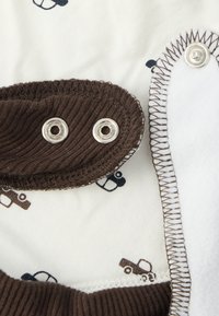 Tejido marrón acanalado con broches de presión plateados, forro blanco suave con patrones de vehículos en azul y marrón, y bordes con costuras contrastadas.