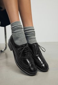 Scarpe Oxford in pelle nera con dettagli brogue, suole piatte e lacci, abbinate a calzini grigi a coste, esposte su una superficie liscia.