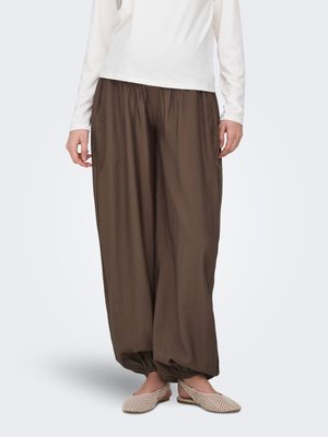 Persona che indossa pantaloni larghi marroni con polsini elastici, una maglia bianca a maniche lunghe e scarpe beige intrecciate senza lacci su uno sfondo uniforme.