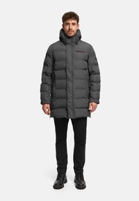 Grijze pufferjas met capuchon, voorzien van een ritssluiting aan de voorkant, een opvallend doorgestikt patroon en een logo-accent op de borst. Draagt over een zwarte broek.