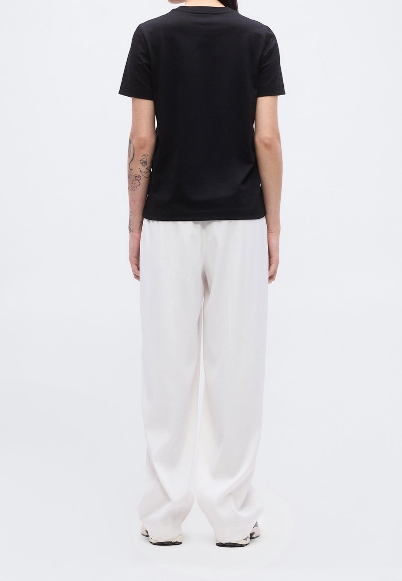 Personne vue de dos portant un t-shirt noir à manches courtes, un pantalon blanc ample et des baskets blanches avec des tatouages visibles sur le bras gauche.