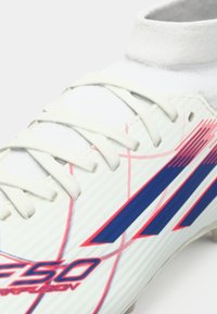 Λευκό ποδοσφαιρικό παπούτσι με ανάγλυφη υφή, διαγώνιες μπλε και κόκκινες ρίγες και λευκά κορδόνια. Σχεδιασμός με ψηλή μοκέτα και εφαρμοστό γιακά για στήριξη.