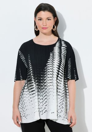 Kvinde iført sort- og hvid plisseret top med abstrakt mønster, øreringe i hoop-stil, stående foran en hvid baggrund.