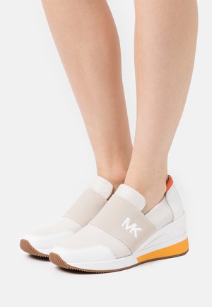 Par hvide slip-on sneakers med beige elastikrem og orange detalje, båret på fødder mod hvid baggrund.