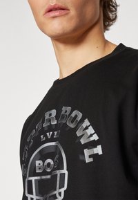 Un primo piano del collo di una persona che indossa una maglietta nera con un design argento del Super Bowl LVII, che presenta un grafico di un pallone da football.