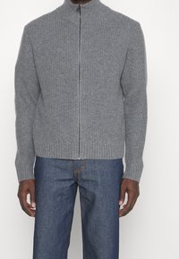 Grå zip-up cardigan gjord av ribbat stickat tyg. Har en hög krage och smala muddar, i kombination med blå denimjeans.