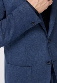 Donkerblauwe getextureerde blazer met een enkele borstzak, twee knopen aan de voorkant en contrasterende zwarte voering bij de manchetten.