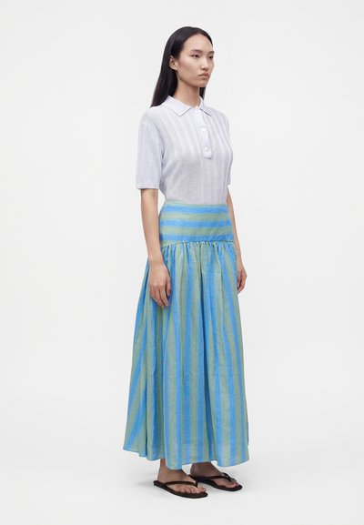 Femme aux longs cheveux foncés portant une chemise blanche à manches courtes boutonnée et une longue jupe rayée bleue et verte avec des sandales noires.
