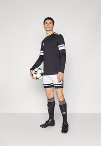 Zwarte lange mouwen shirt met witte strepen, witte shorts met zwarte strepen, zwarte sokken, zwarte voetbalschoenen en een gestructureerde voetbal.