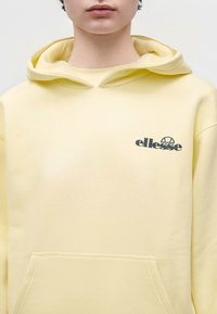 Personne portant un sweat à capuche ellesse jaune pâle avec poche avant, logo sur la poitrine et capuche reposant sur les épaules sur un fond uni.
