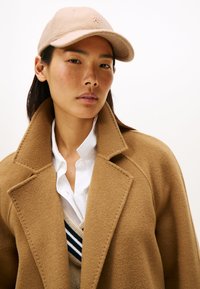 Abrigo de lana beige con solapas anchas, superpuesto a una camisa blanca y un suéter beige a rayas. Gorra rosa con un pequeño logo en la parte frontal.