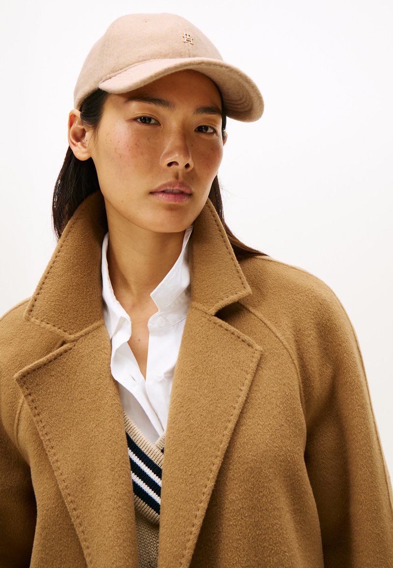 Abrigo de lana beige con solapas anchas, superpuesto a una camisa blanca y un suéter beige a rayas. Gorra rosa con un pequeño logo en la parte frontal.