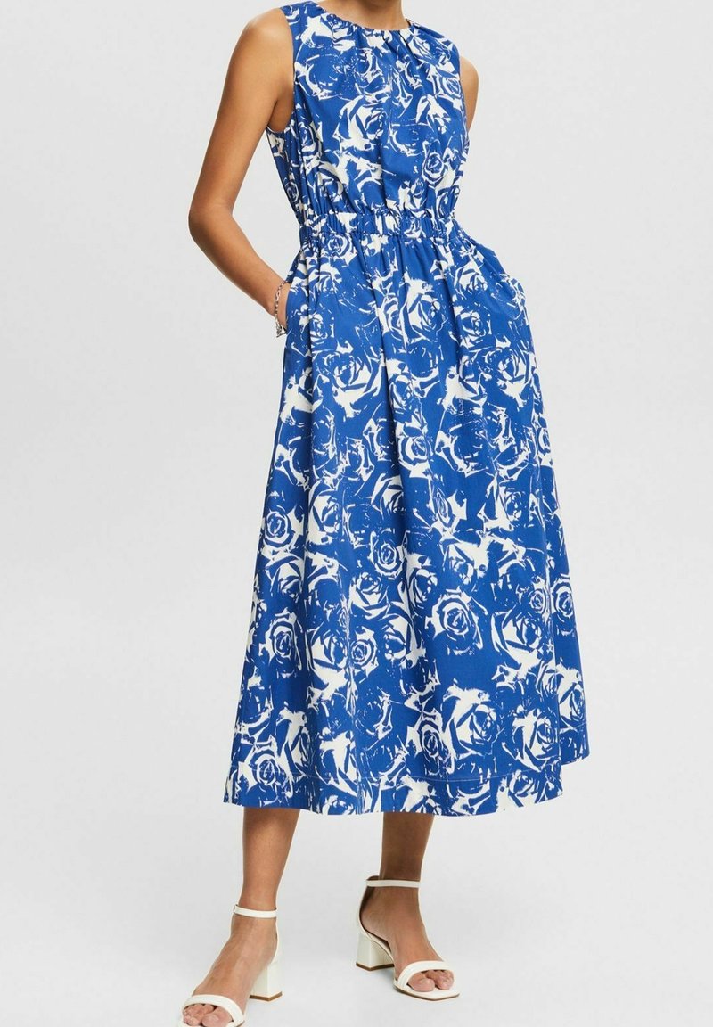 Robe midi bleu sans manches avec motif floral abstrait blanc, taille élastique, poches latérales, portée avec des sandales blanches à talons blocs.