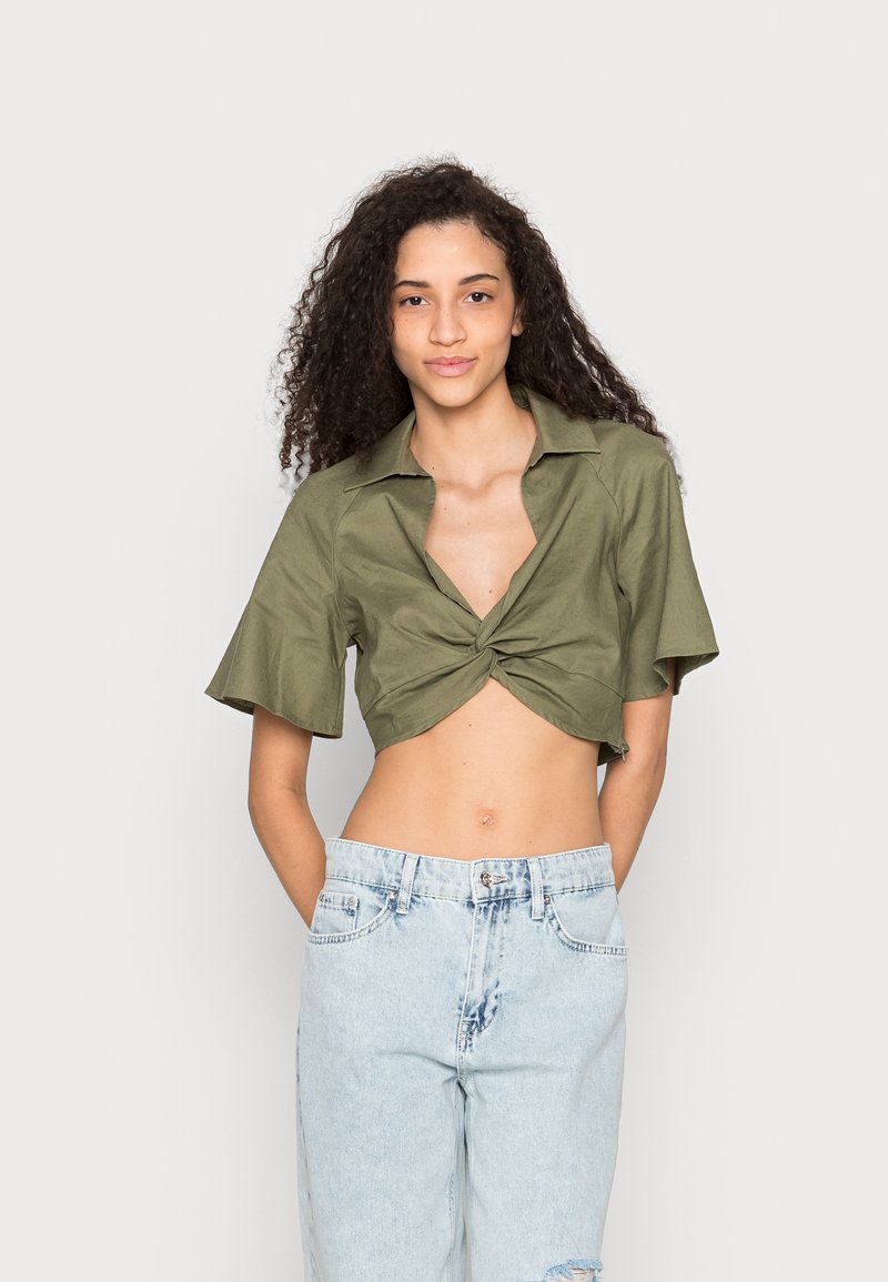 Camisa verde oliva corta com frente atada, mangas curtas e gola, combinada com calças de ganga desbotadas e rasgadas.