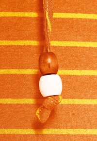 Oranssin kangas, jossa on vaakasuoria keltaisia raitoja ja kudottu nyöri. Kaksi helmeä, yksi puinen ja yksi valkoinen, korostaa nyöriä.