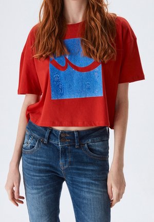 Vrouw met lang rood haar, die een rood cropped t-shirt met een blauw-rood abstract ontwerp draagt en blauwe jeans met dubbele knoop.