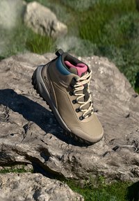 Bota de senderismo beige con una superficie texturizada, acentos en rosa y azul, suela resistente y seis ojales de metal para los cordones. Colocada sobre una superficie rocosa.