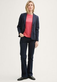 Blazer marine sur un T-shirt corail, associé à un jean évasé en denim foncé et des mocassins noirs. Le mannequin se tient avec une main dans la poche, sur un fond neutre.