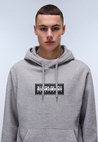 Felpa grigia con cappuccio e cordoncino, grande tasca frontale e patch logo in bianco e nero sul petto. Tessuto morbido e vestibilità comoda.