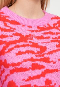 Knit sweater featuring a vibrant red and pink zigzag pattern, with a ribbed pink neckline and soft texture. Close-up view.