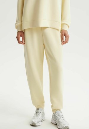 Osoba ubrana w kremowe spodnie dresowe i pasującą bluzę, stojąca, widoczne białe i szare buty sportowe.