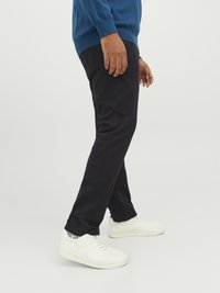 Svarta tajta byxor med en slät yta, kombinerade med vita sneakers. Modellen bär en blå tröja och visas från sidan.