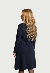 Donna con lunghi capelli biondi ondulati, che indossa un abito lungo nero, in piedi di lato contro uno sfondo chiaro e semplice.