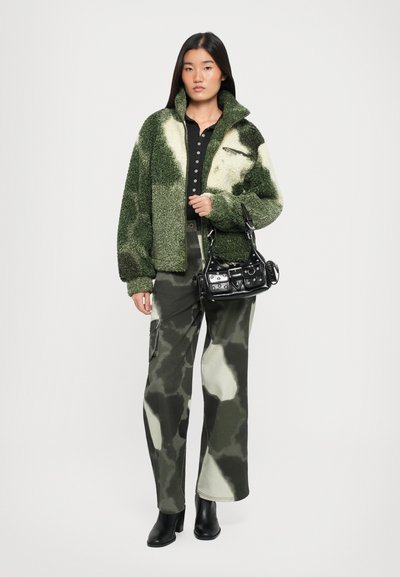 Ylikokoiset vihreä ja helmenvalkoinen fleece-takki, musta nappipaita, camo cargo-housut ja pieni musta laukku. Mallilla on päällään mustat nilkkurit.