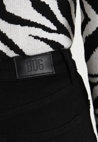 Vaqueros de mezclilla negra con un tejido texturizado, una etiqueta de cuero de la marca "BDG" y detalles de costura visibles.