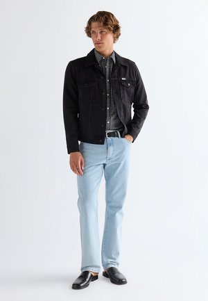 Mann som står med hånden i lomma, iført svart jakke, svart-hvitt rutete skjorte, lyseblå jeans og svarte slip-on sko.