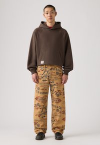 Sudadera marrón con capucha y bolsillo frontal, combinada con pantalones beige holgados con diseños de impresión vintage y un corte relajado. Zapatos negros.