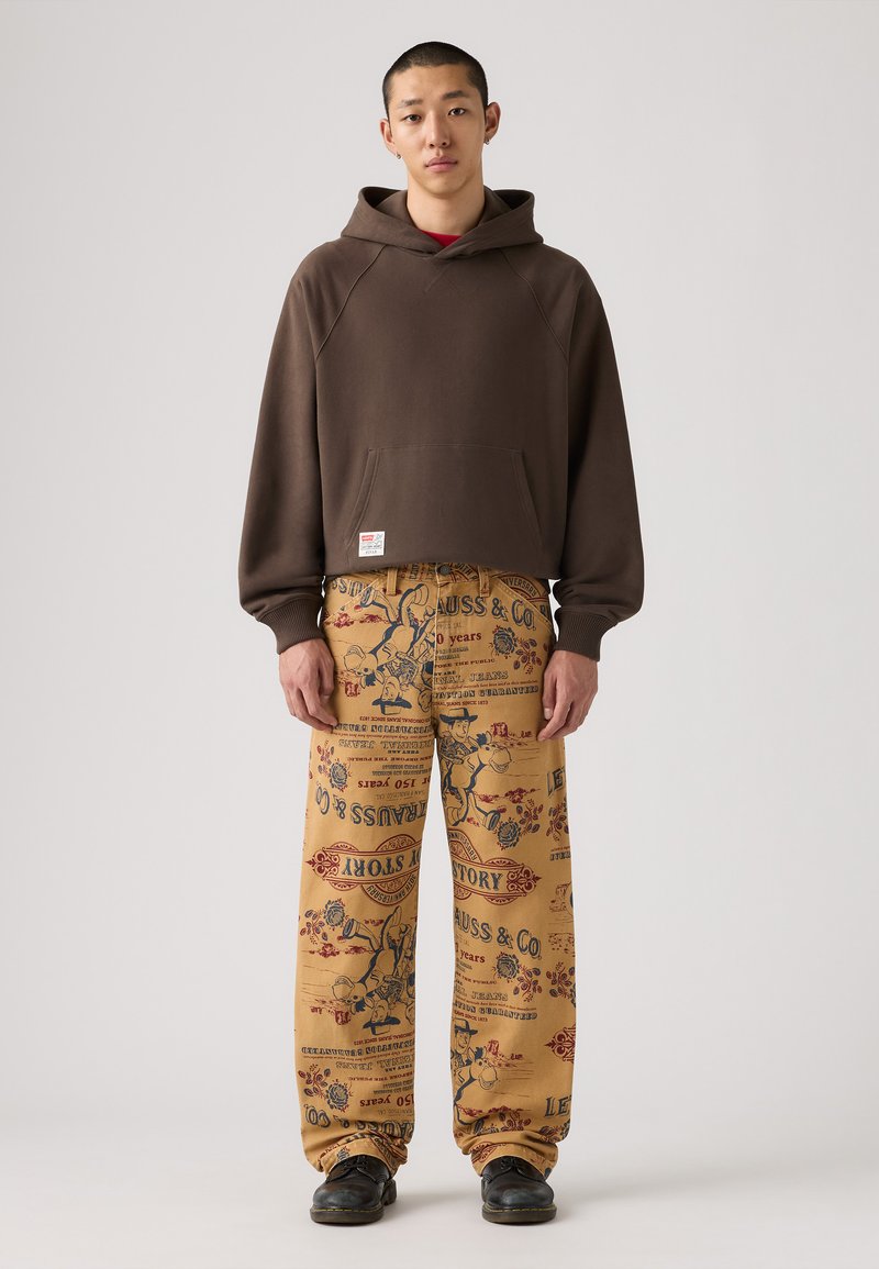 Sudadera marrón con capucha y bolsillo frontal, combinada con pantalones beige holgados con diseños de impresión vintage y un corte relajado. Zapatos negros.