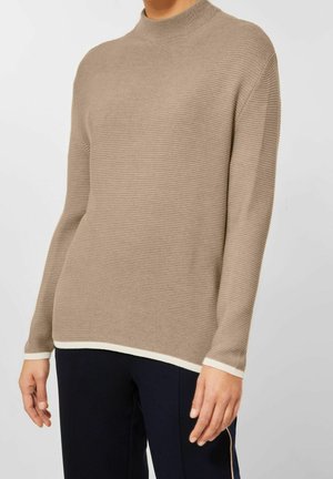 Personne portant un pull beige côtelé à manches longues avec finitions blanches aux poignets et à l’ourlet, assorti à un pantalon foncé sur un fond uni.