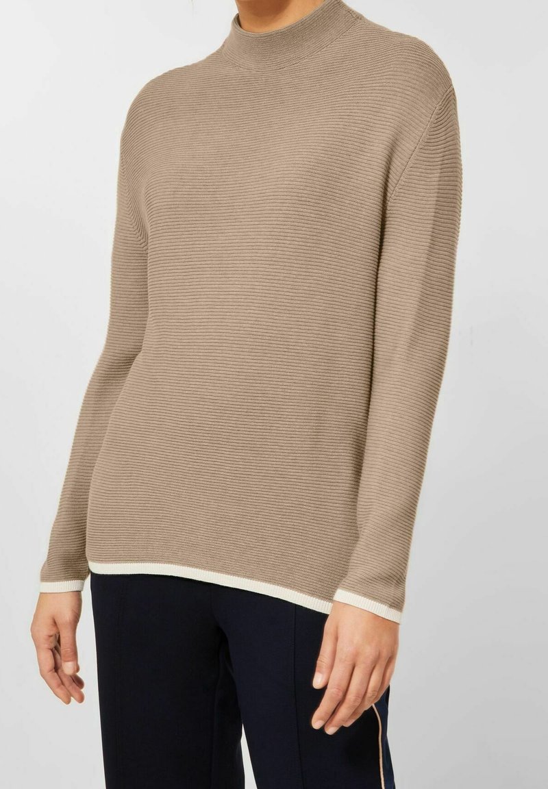 Personne portant un pull beige côtelé à manches longues avec finitions blanches aux poignets et à l’ourlet, assorti à un pantalon foncé sur un fond uni.