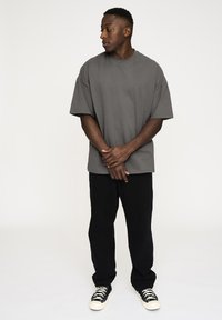 Grå oversized T-shirt med korte ærmer, parret med sorte bukser og sort-hvide sneakers, stående på en hvid baggrund.
