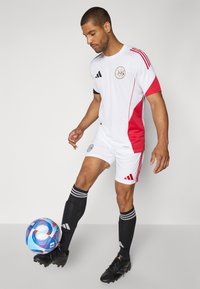 Maillot de football blanc et rouge avec un logo Adidas, arborant des manches courtes, des accents noirs et des chaussettes noires jusqu'aux genoux, accompagné d'un ballon coloré.