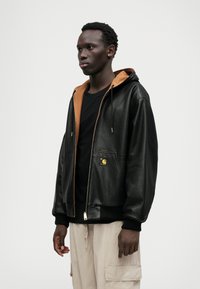 Μαύρη δερμάτινη κουκούλα με καφέ εσωτερικό, διαθέτει μπροστινό φερμουάρ, δύο τσέπες και ένα μικρό λογότυπο, σε συνδυασμό με μαύρο πουκάμισο και ανοιχτόχρωμο παντελόνι.