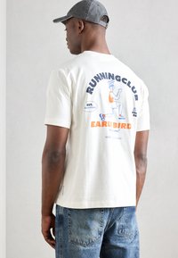 Λευκό βαμβακερό μπλουζάκι με γραφικό "RUNNINGCLUB" σε ναυτικό μπλε και πορτοκαλί στην πλάτη, συνδυασμένο με ανοιχτό μπλε τζιν. Απλός σχεδιασμός.