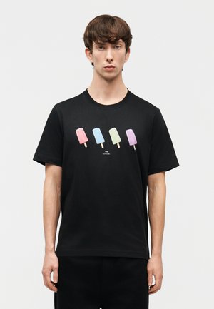 Νεαρός άνδρας που φοράει μαύρο μπλουζάκι με τέσσερα παγωτίνια σε παστέλ χρώματα σχεδιασμένα στο ύψος του στήθους, στέκεται μπροστά σε απλό φόντο.