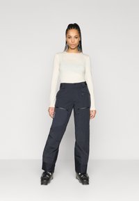 Donkerblauwe skibroek met cargozakken, verstelbare taille en met ritsen. Draagt samen met een licht shirt met lange mouwen en skischoenen.