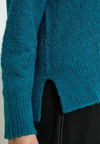 Teal wool sweater featuring a textured finish, ribbed cuffs, and a side slit for added detail, displayed with a black garment underneath.