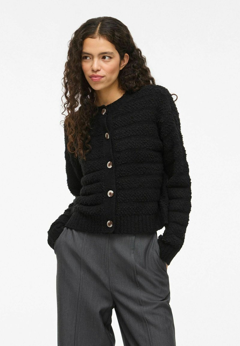 Zwarte gebreide cardigan met knoopsluitingen, ronde halslijn en geribbelde manchetten. Gepaard met grijze high-waisted broeken voor een gelaagd uiterlijk.