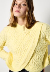 Żółty sweter w splot warkoczowy z wysokim kołnierzem i ukośnym elementem wzoru. Miękka faktura, widoczny wzór dzianiny oraz prążkowane wykończenia na dole.