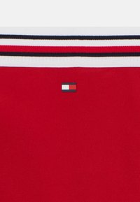 Rød stof med en glat tekstur, der har et hvidt og sortstribet design øverst. Inkluderer et lille logo-patch i blå, hvid og rød.