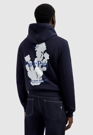 Man draagt een marineblauwe hoodie met een witte bloemenafbeelding en de tekst "Pure Path" op de achterkant, gecombineerd met donkerblauwe spijkerbroek, en staat met de rug naar voren.