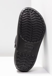 Semelle de Crocs noire montrant un motif de bande de roulement avec textures en zigzag et points, marquages de taille "M 4" et "W 6" visibles sur une surface blanche.