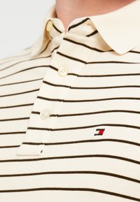 Μπεζ ριγέ πόλο πουκάμισο με στενές σκούρες ρίγες, τρία κουμπιά στο μπροστινό μέρος και κεντημένο λογότυπο στο στήθος. Υφή υφάσματος.